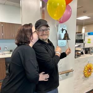 Hispanic couple both dressed in black se is kissing him as he rings the bell and holds balloons