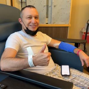 middle age white man with short hair, a black mask pulled down, a blue sleeve on his arm giving a thumbs up wearing white shirt with plaid shorts
