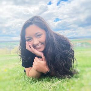 smiling young hispanic girl with long dark hair lying in the grass head propped on hand wearing black shirt and watch