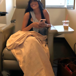 young ethnic girl sitting in a hospital chair wearing a yellow ball cap and a black sleevelss tshirt holding a pink phone with a blanket on her lap