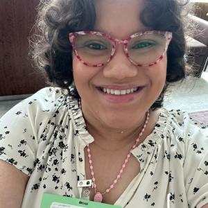 young hispanic woman with dark curly hair and red and white glasses wearing a white shirt with black flowers