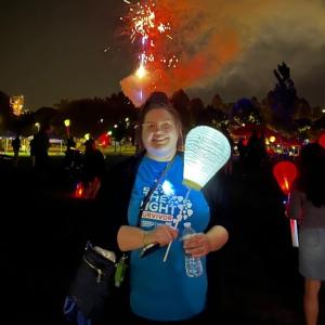 young white girl with her hair pulled up with glasses wearing a black jacket and blue tshirt carrying a white balloon at LTN with fireworks in the background