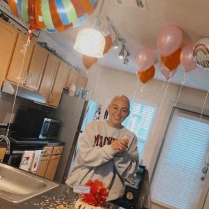 young white woman bald with a nose ring wearing a sweatshirt and standing in a kitchen with balloons and a bundt cake