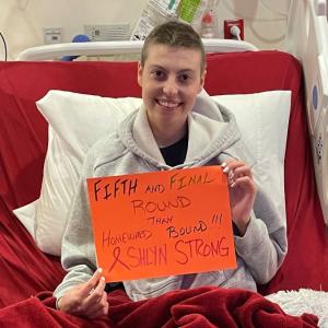 young white woman in a hospital bed with short hair wearing a gray hoodie and holding an orange sign about fifth round of chemo