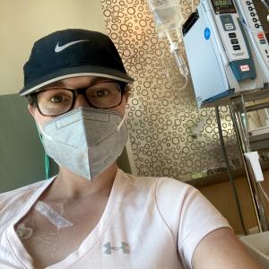 young white woman with short hair wearing a blue ball cap, brown glasses, a face mask and a pink Underarmor shirt