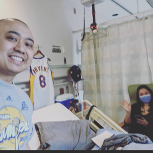 Nino Asian man in hospital room wearing blue tshirt standing by a Kobe Bryant jersey with his wife sitting in a chair waving and wearing a blue mask and dark shirt
