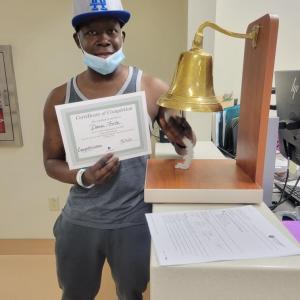 middle aged black man wearing LA ballcap and gray and white tank top and gray sweat pants holding a certificate and ringing a bell.
