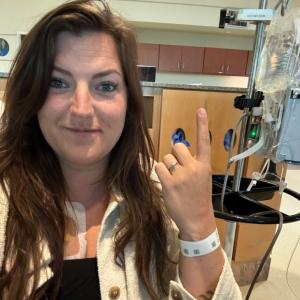 young white woman with long brown hair wearing a black shirt and white jacket sitting in a hospital room holding up her pointer finger