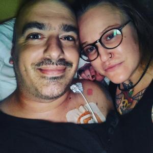 younger white man with bushy eyebrows and mustache wearing a black shirt with a port lying next young white woman with black glasses and a black nose ring with a tattoo on her finger and neck