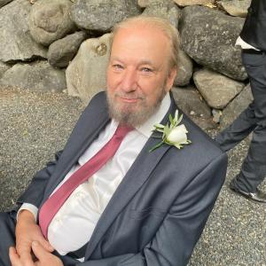 Gary older white man with blue eyes and thinning hair and gray beard and mustache leaning on rocks wearing a blue suite, mauve tie and a rose boutonaire