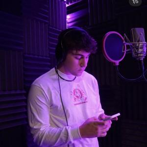 Marko young white male with curly dark hair in a recording studio wearing headphones and a puka shell neclklace and a white long sleeve tshirt with a pink logo also holding a phone