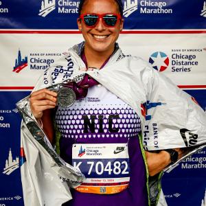 Nicolas smiling middle aged hispanic woman with dark hair pulled up in a bun wearing orangish sunglasses and a Team In Training Shirt holding a medallion in front of a Chicago Marathon banner.jpg