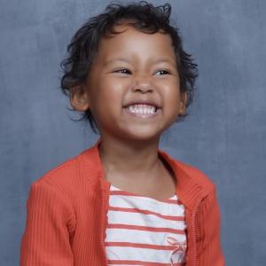 Sofia school picture of pre-K black girl with short curly hair and a big smile wearing and orange and white striped top under an orange sweater with a zipper