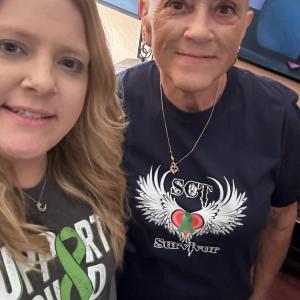 Susanne middle aged white woman with a bald head wearing hoop earrings and a heart necklace and a SCT Survivor t-shirt standing next to a young woman with a nose ring and Support Squad t-shirt