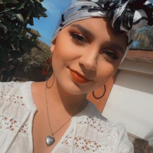 Yesenia young hispanic woman with blue and black headscarf on hoop earrings and a heart necklace wearing a white lacey blouse