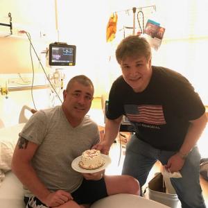 Greg two middle aged white men, one sitting on a hospital bed wearing a grey t-shirt and black shorts the other wearing a flag shirt and jeans both holding a cake