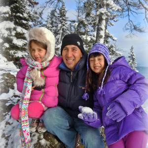 Joaquin middle aged hispanic man wearing navy beanie and ski jacket with two hispanic girls one in a pink jacket and one in a purple jacket holding a snowball in front of snow covered trees