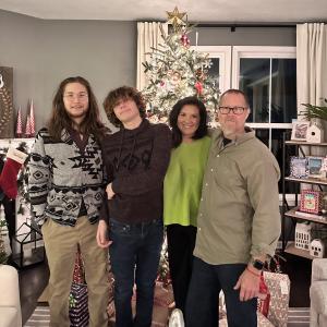 John William middle aged white man wearing glasses and a gray shirt standing in front of Christmas tree with his wife in a green sweater and two sons