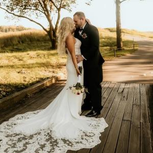 Kayla young woman with long blond hair in a white spagetti strap wedding dress holding a bouquet with arm around a young white man with beard and mustache.
