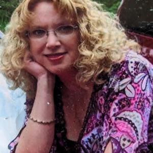 middle aged white woman with curly blonde hair and glasses with her chin resting on her hand wearing a pink and purple print shirt
