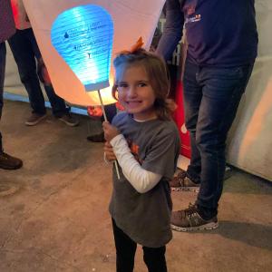 young smiling white girl in a gray shirt over a white long sleeved shirt with and orange bow in her hair and holding a LTN balloon