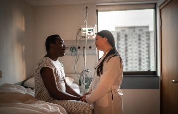 An African-American patient holds the hand and speaks with a doctor of Asian descent in a hospital room