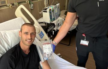 Young man in black t-shirt with leukemia in a hospital bed with a medical professional standing next to him wearing a mask