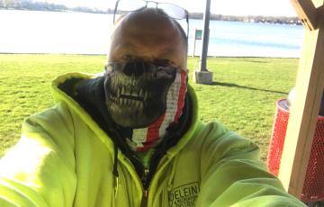 balding white man with sunglasses on his head wearing a monster mask and yellow hoodie in front of water