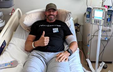 middle aged white man in a ball cap with a scruffy beard and mustached wearing a black t-shirt lying in a hospital bed giving a thumbs up