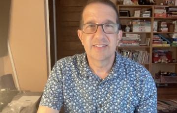 middle aged white man wearing glasses and a blue print shirt sitting in front of a bookcase