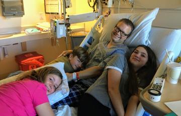 middle aged white man wearing glasses and a hospital gown sitting on a hospital bed with his son and two daughers