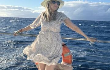 middle aged white woman wearing a straw hat sunglasses and a floral dress leaning on a boat's railing