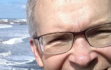 older white man with brown framed glasses plaid shirt and black jacket and big smile standing in front of the ocean