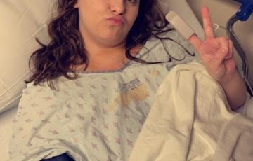 young woman with long brown hair wearing hosptial gown puckering her mouth and giving a peace sign lying in a hospital bed