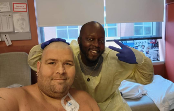 younger white male with shower cap on scruffy beard and no shirt sitting in front of black male medical tech giving a peace sign