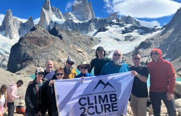 a group of nine people standing at the top of a mountain holding a Climb2Cure banner