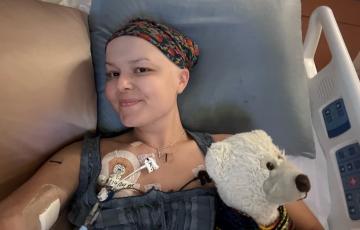 young white woman wearing a head scarf and gray top lying in a hospital bed with a stuff white bear