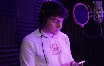 Marko young white male with curly dark hair in a recording studio wearing headphones and a puka shell neclklace and a white long sleeve tshirt with a pink logo also holding a phone