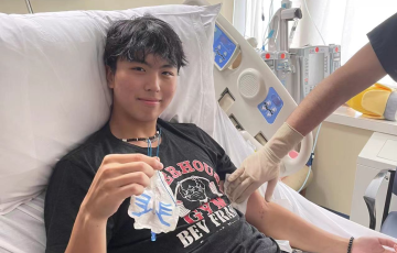 Kevin young asian young man with black hair wearing a puka shell necklace and black gym tshirt and shorts while laying in a hospital bed