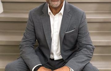 Arturo Pierre middle aged black man bald with mustache and beard wearing black glasses and a grey suite and white shirt sitting on steps